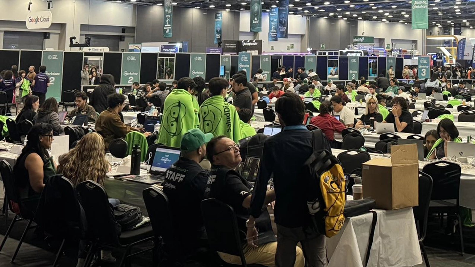 Sala del Talent Hackathon con equipos trabajando en laptops; al centro, participantes con mantas verdes de Salud Digna y personal de staff